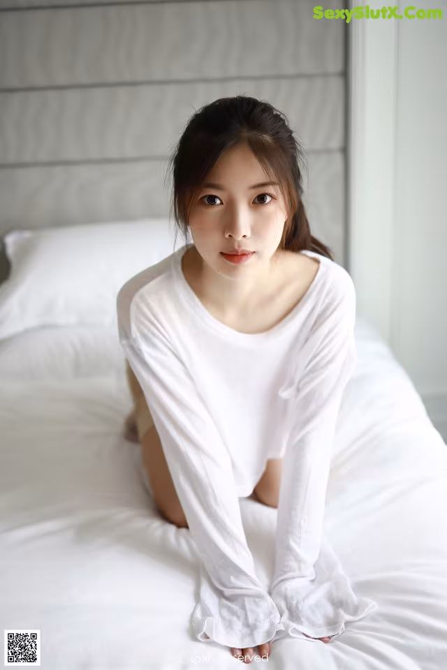 A woman sitting on a bed in a white shirt.