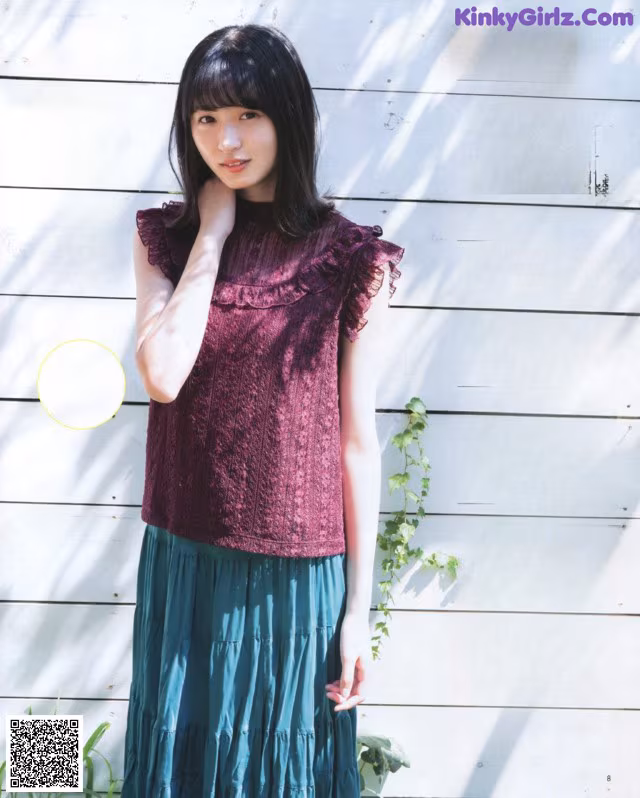 A woman standing in front of a white wall wearing a purple top.